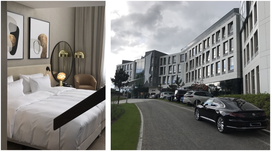 Zdjęcie przedstawia wnętrze pokoju hotelowego z widokiem na budynek hotelu na zewnątrz. W pokoju znajduje się duże łóżko z białymi poduszkami i kołdrą, obrazy na ścianie, okrągłe lustro i lampa.