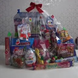 Świąteczny zestaw upominkowy zapakowany w przezroczystą folię z czerwoną kokardą, zawierający słodycze, przekąski i zabawki dla dzieci, na tle ściany w geometryczne wzory.