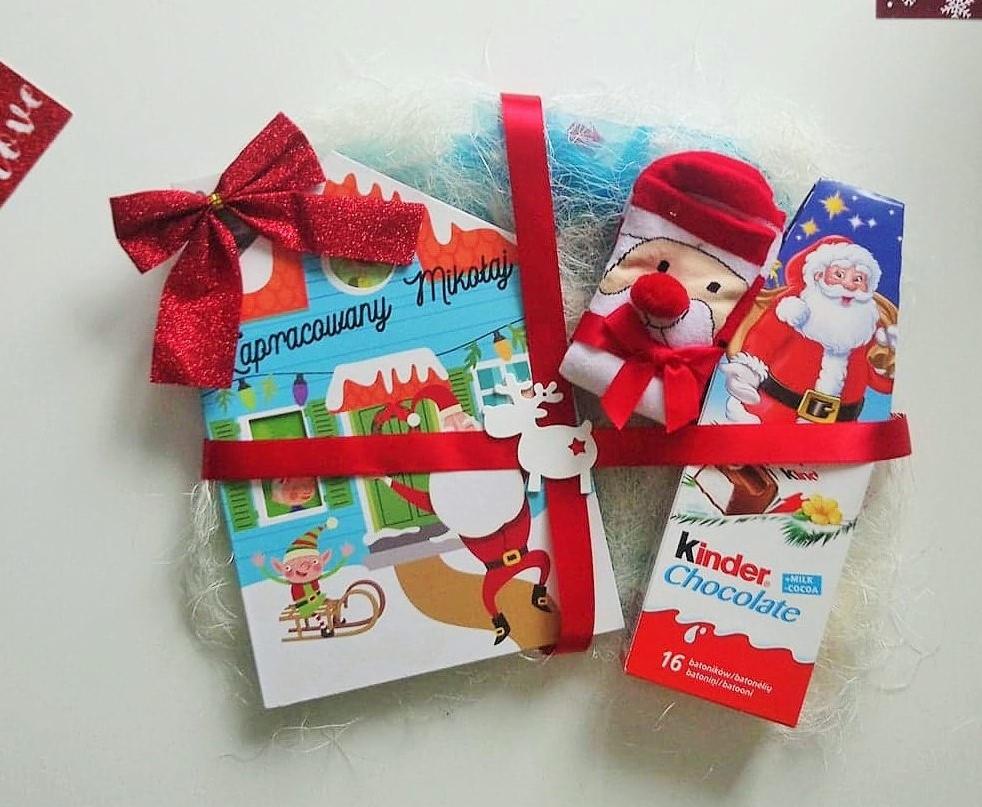 Świąteczny zestaw prezentowy z książką 'Zapracowany Mikołaj', skarpetkami z Mikołajem, czekoladkami Kinder, przewiązany czerwoną wstążką i ozdobiony reniferem.
