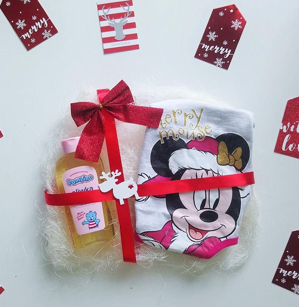 Zestaw prezentowy dla dziecka z oliwką Bambino i ubrankiem z motywem Myszki Minnie, przewiązany czerwoną wstążką z kokardą i drewnianym reniferem, na białym tle z dekoracyjnymi etykietami.
