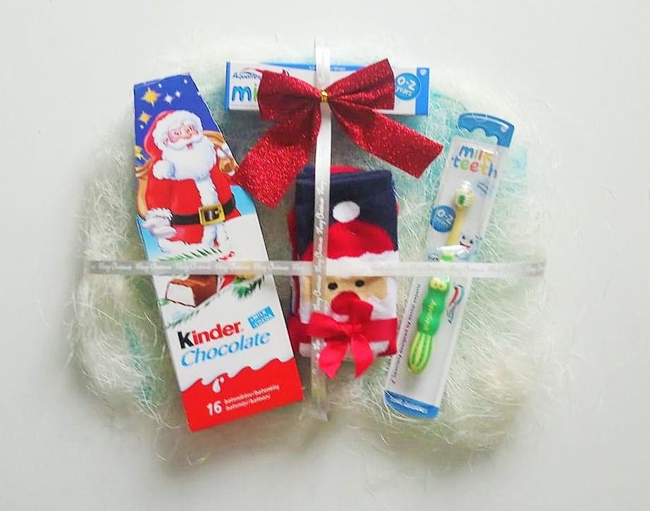 Świąteczny kosz prezentowy dla dziecka z czekoladą Kinder, szczoteczkami do zębów dla niemowląt i skarpetką z motywem Świętego Mikołaja, przewiązany błyszczącą czerwoną kokardą i przezroczystą...