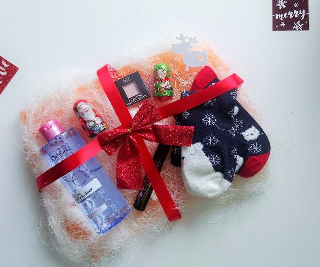 Świąteczny zestaw upominkowy z kosmetykami L'Oreal, skarpetkami w śnieżynki, czekoladowymi figurkami i cieniem do powiek, przewiązany czerwoną, brokatową wstążką na tle wypełniacza z włókien.