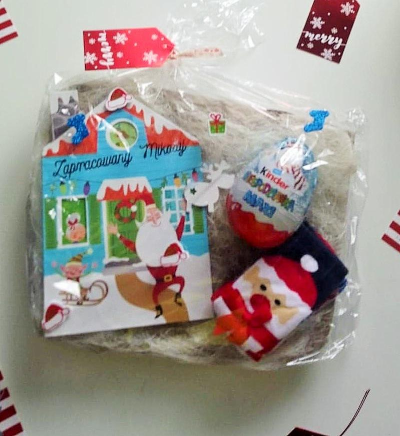 Świąteczny upominek w folii: kartka z Mikołajem przed domkiem, jajko Kinder Niespodzianka Maxi i skarpetki z motywem Mikołaja, wypełnienie z włókna, dekoracyjne naklejki i zawieszka Merry.