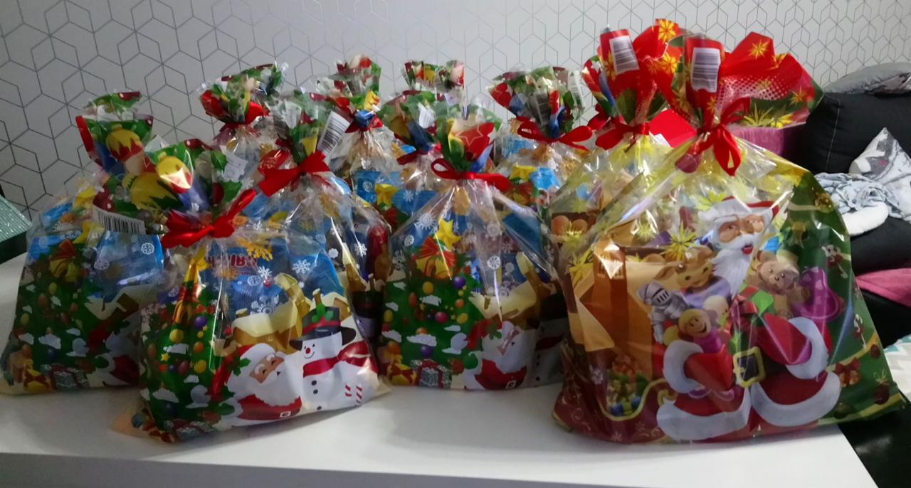 Spora grupa zapakowanych prezentów w przezroczystych torbach z motywami świątecznymi, przewiązanych czerwonymi wstążkami, ustawionych na białym stole.
