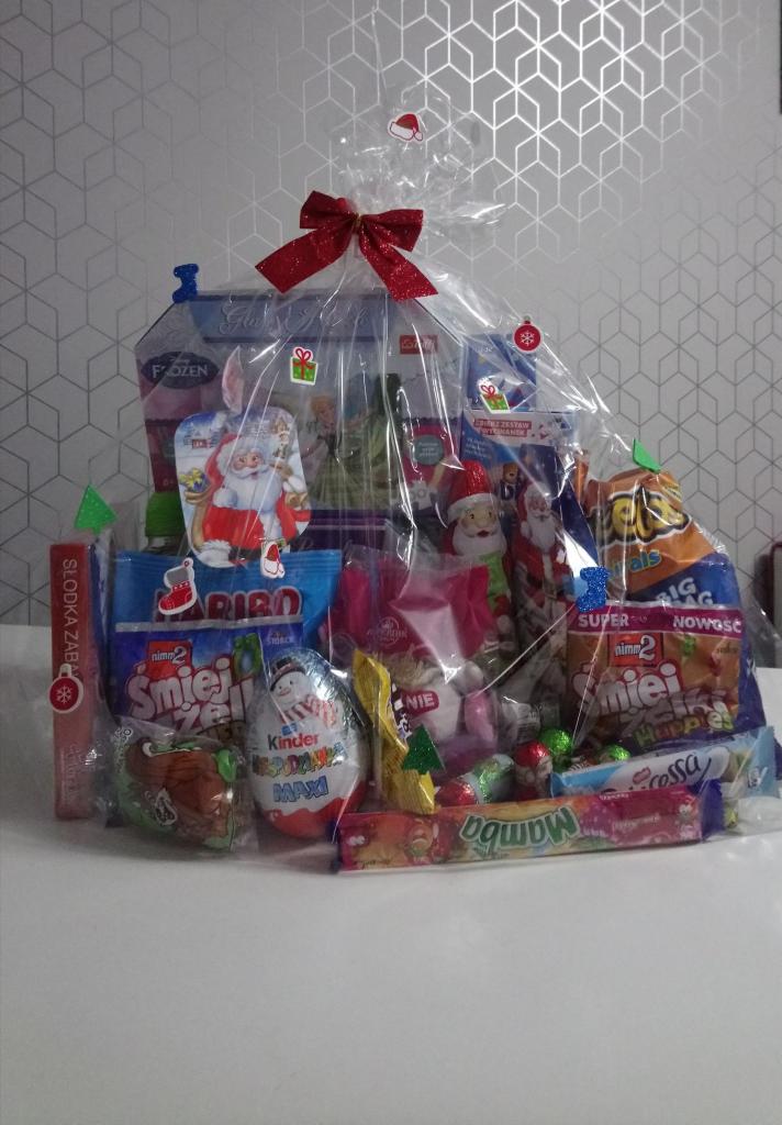 Świąteczny zestaw upominkowy zapakowany w przezroczystą folię z czerwoną kokardą, zawierający słodycze, przekąski i zabawki dla dzieci, na tle ściany w geometryczne wzory.