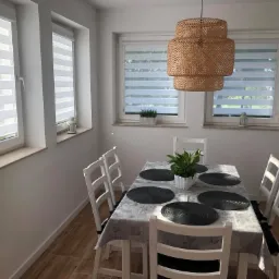 Jasne wnętrze jadalni z trzema oknami wyposażonymi w rolety poziome dzień i noc, nad stołem wisi pleciona lampa, a na parapecie ozdobne lampiony.