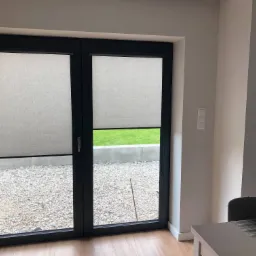 Zamknięte, jasne rolety wewnętrzne w czarnej ramie okiennej, widok na żwirowy ogród i trawnik.