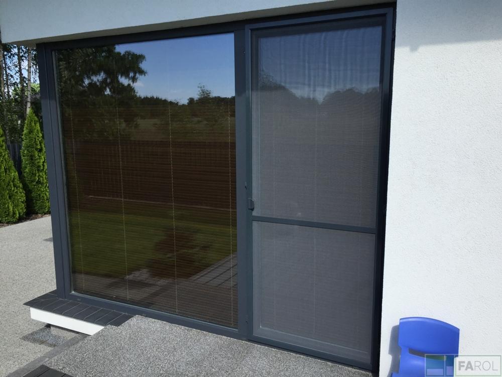 Szara rama okienna z moskitierą po prawej stronie i brązowymi żaluzjami widocznymi przez szybę po lewej, na tle białej ściany i zieleni ogrodu.