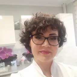 Portret kobiety w okularach z delikatnym makijażem ust i brwi w salonie kosmetycznym, w tle dekoracja z motywem kwiatowym i szafki.