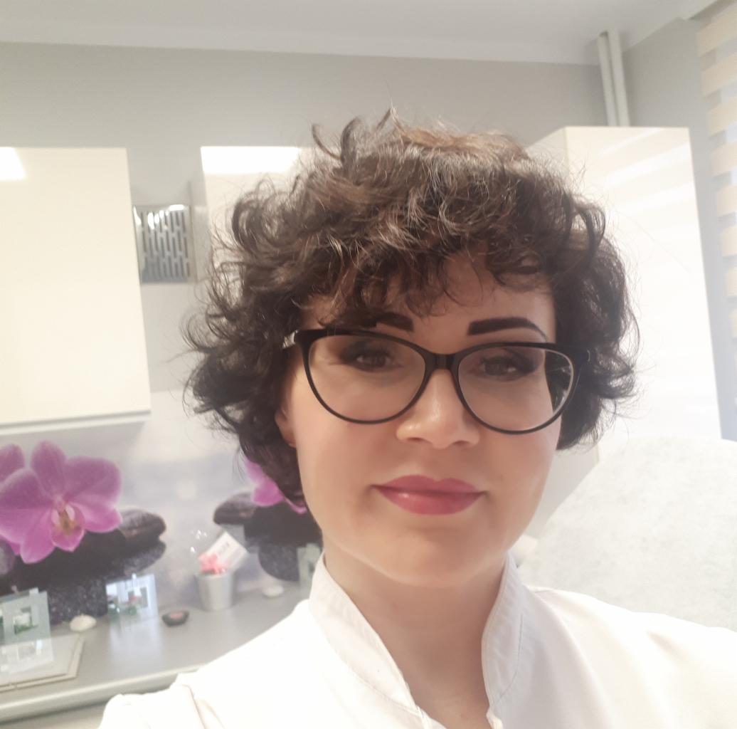 Portret kobiety w okularach z delikatnym makijażem ust i brwi w salonie kosmetycznym, w tle dekoracja z motywem kwiatowym i szafki.