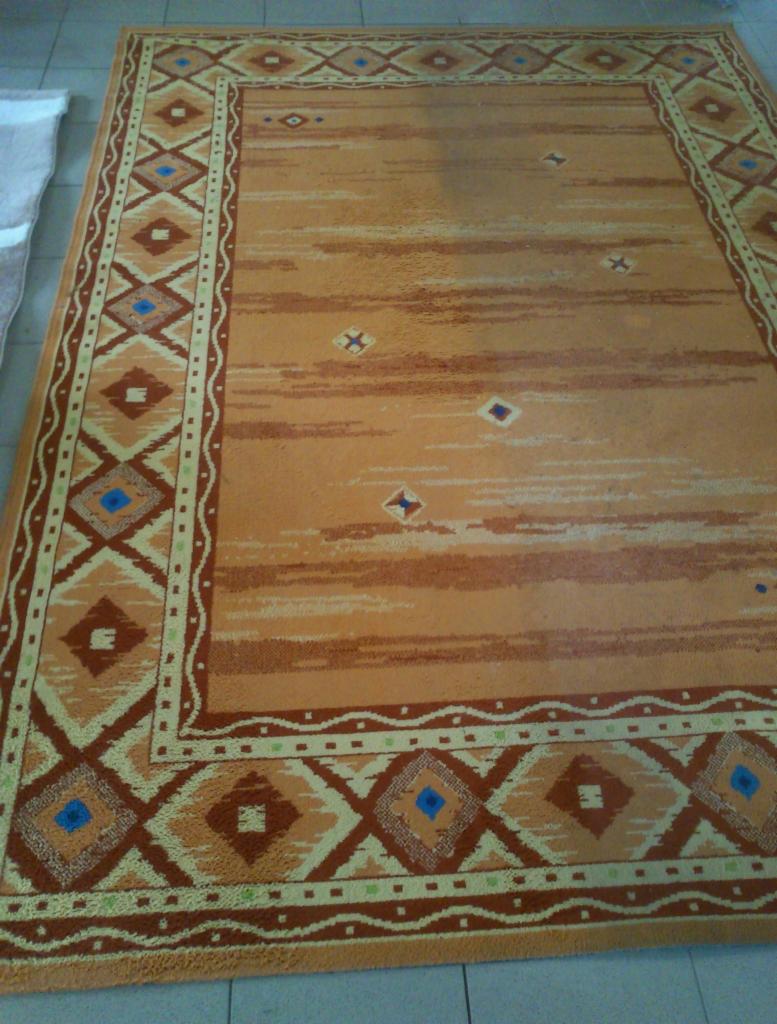 Beżowy dywan z geometrycznym wzorem i brązowymi akcentami leży na podłodze z płytek.