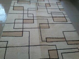 Beżowy dywan z geometrycznym wzorem z prostokątów i linii w kolorze czarnym i beżowym, leżący na brązowej podłodze z płytek.