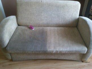 Dwukolorowa kanapa po częściowym praniu, widoczna różnica między wyczyszczoną a brudną powierzchnią, na oparciu leży fioletowa gumka do włosów.