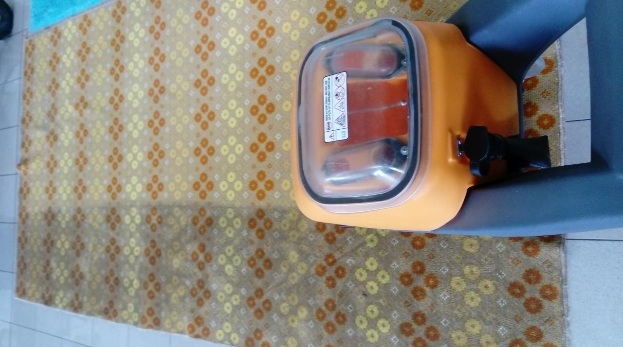 Pomarańczowe urządzenie do czyszczenia dywanów z przezroczystym zbiornikiem, stojące na wzorzystym dywanie.