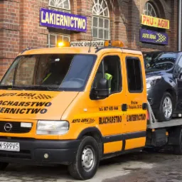 Pomarańczowa laweta z napisem 'Blacharstwo Lakiernictwo' przewozi szary samochód osobowy na tle ceglanego budynku z szyldami reklamowymi.