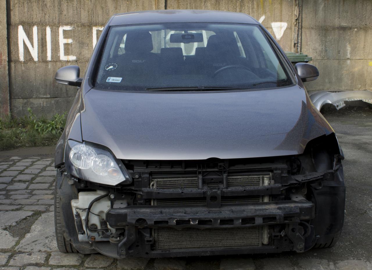 Uszkodzony przód szarego samochodu marki Golf, widoczny na tle betonowego muru z białym napisem 'NIE', brakujący element karoserii po stronie kierowcy, odsłonięty chłodnica.