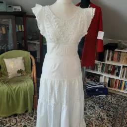 Biała, długa suknia z ażurowymi wstawkami, prezentowana na manekinie w domowym otoczeniu z dywanem w orientalne wzory, krzesłem z zielonym pledem i regałem z książkami w tle.