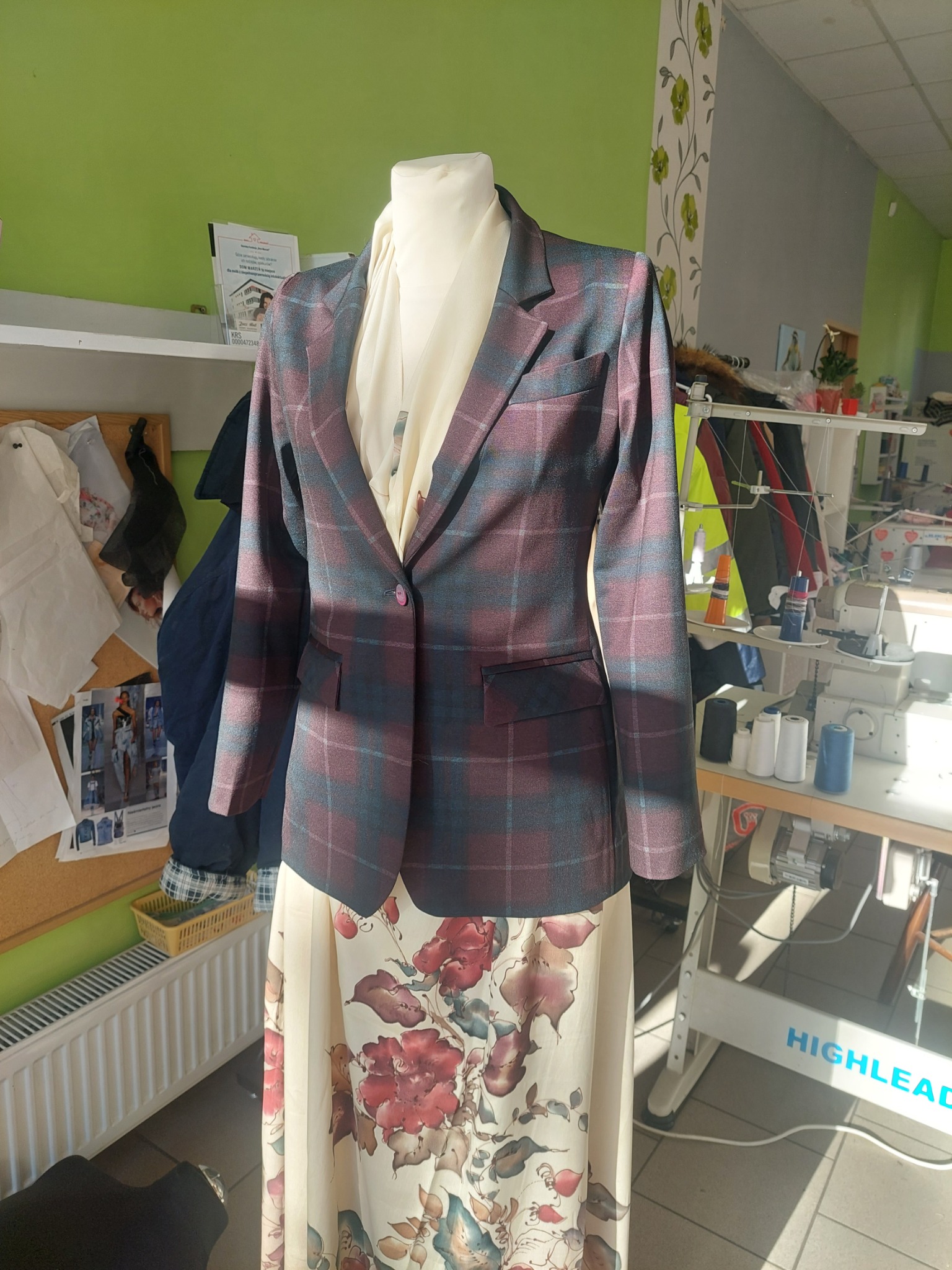 Manekin w pracowni krawieckiej prezentuje uszyty na miarę, bordowo-granatowy żakiet w kratę z jednym guzikiem, zestawiony z kwiecistą spódnicą.