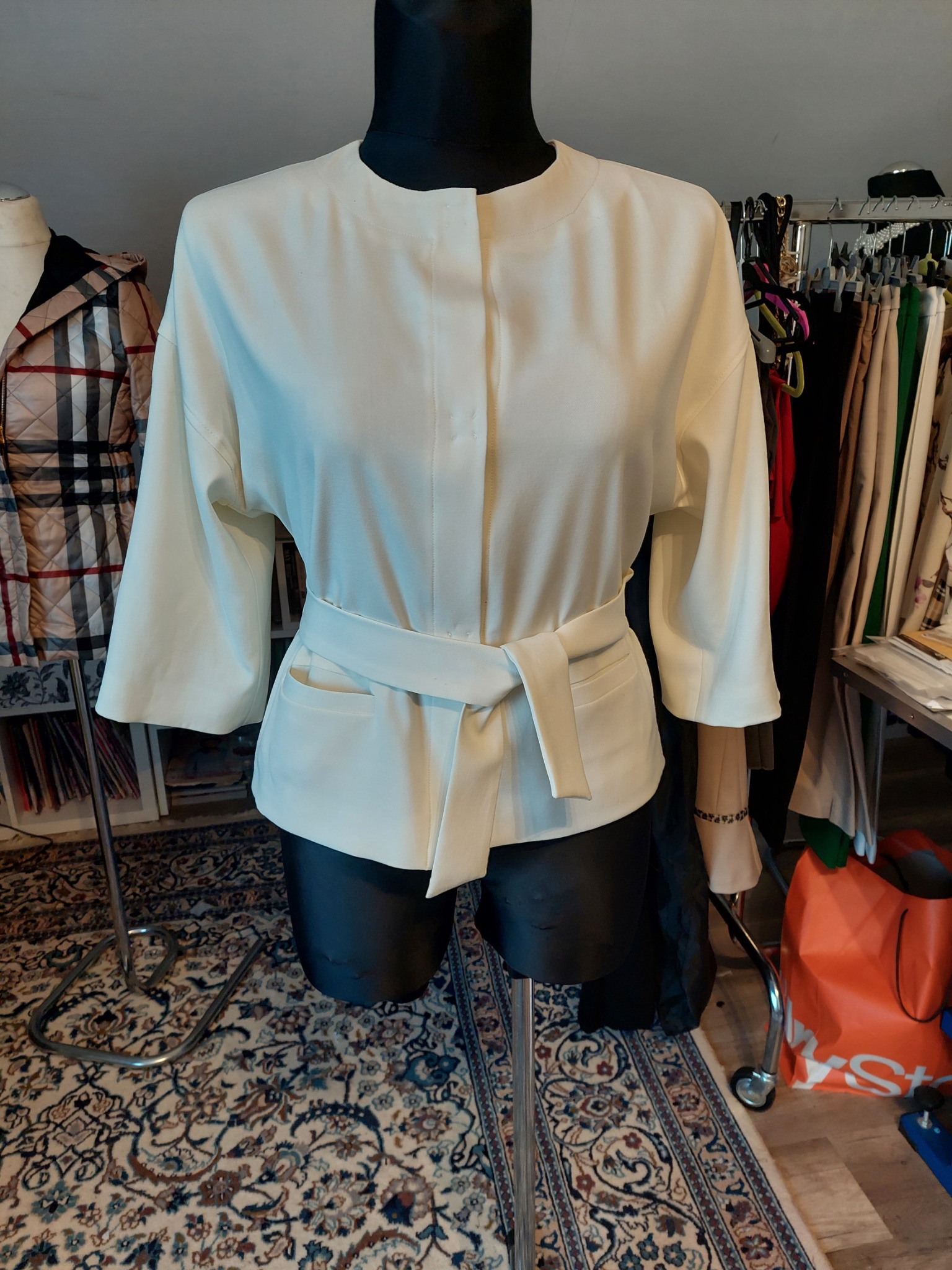 Kremowa bluzka z szerokimi rękawami i wiązanym paskiem prezentowana na czarnym manekinie w pracowni krawieckiej, na tle wieszaków z ubraniami i wzorzystego dywanu.