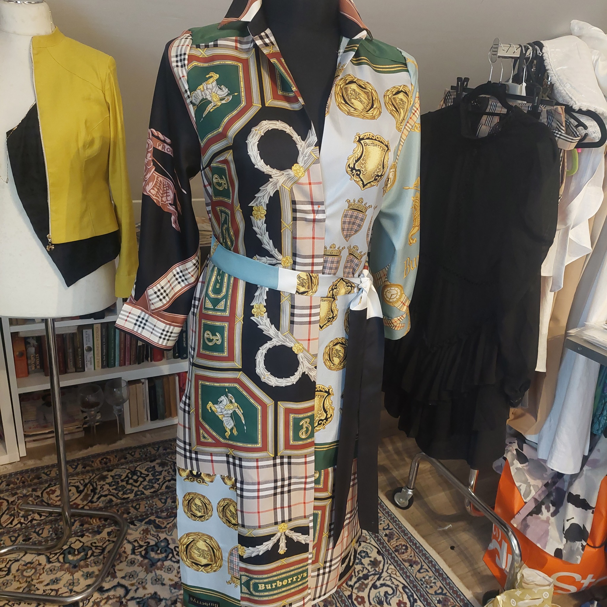Sukienka koszulowa na manekinie z wzorem w heraldyczne motywy i kratę Burberry, z paskiem w talii, w tle widać inne ubrania i regał z książkami.