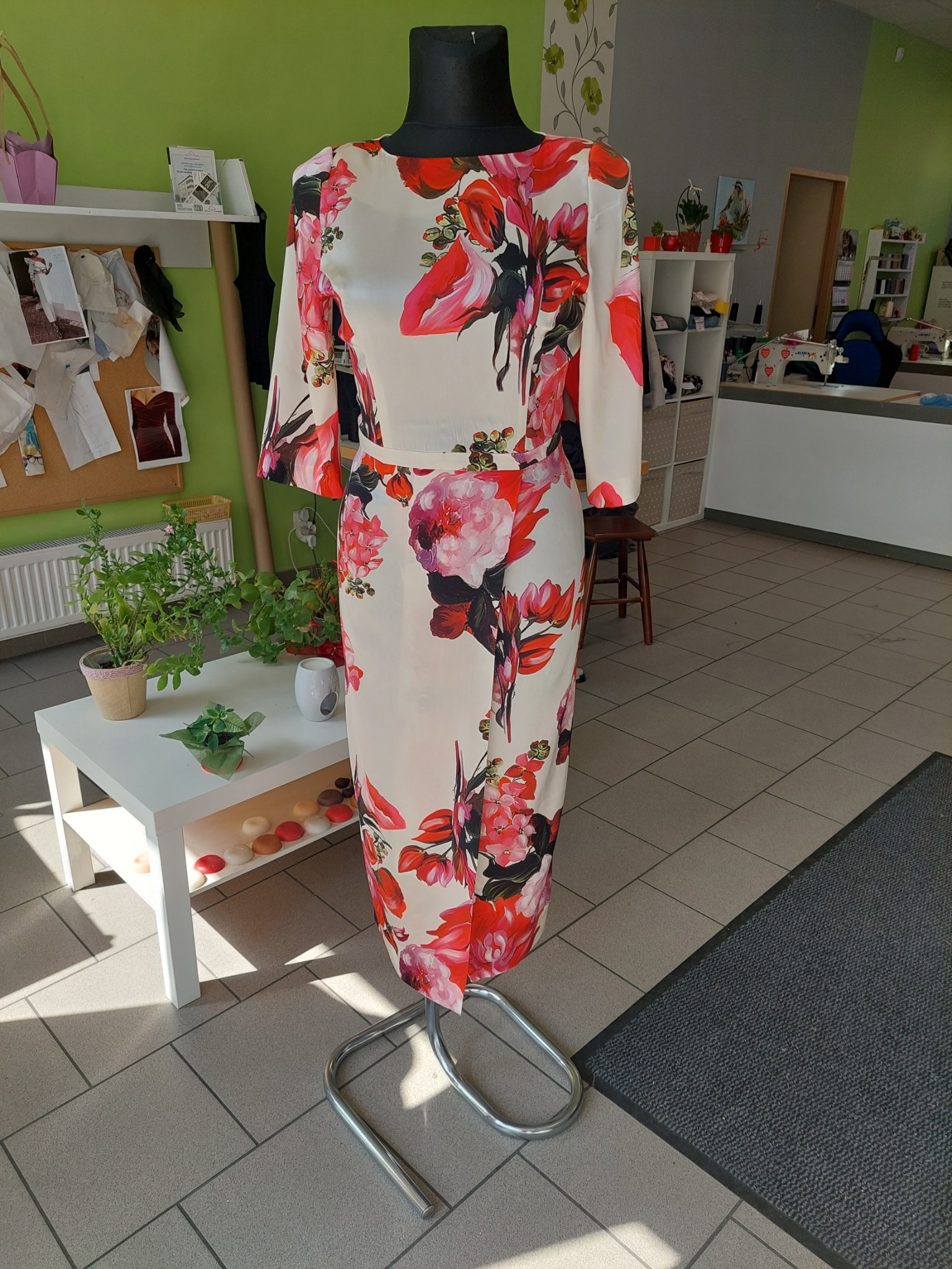 Sukienka z kwiecistym wzorem na manekinie w pracowni krawieckiej, widoczne biurko i akcesoria.