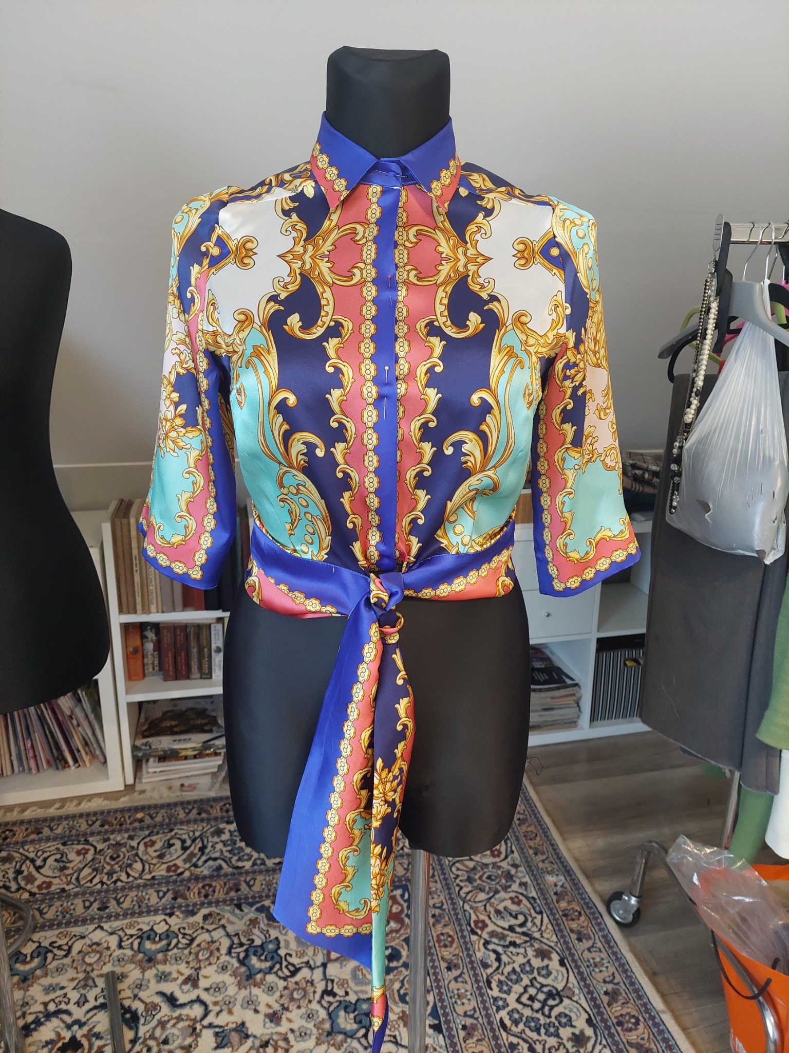 Bluzka z barokowym wzorem, zawiązywana w talii, prezentowana na czarnym manekinie krawieckim z widocznymi szpilkami.