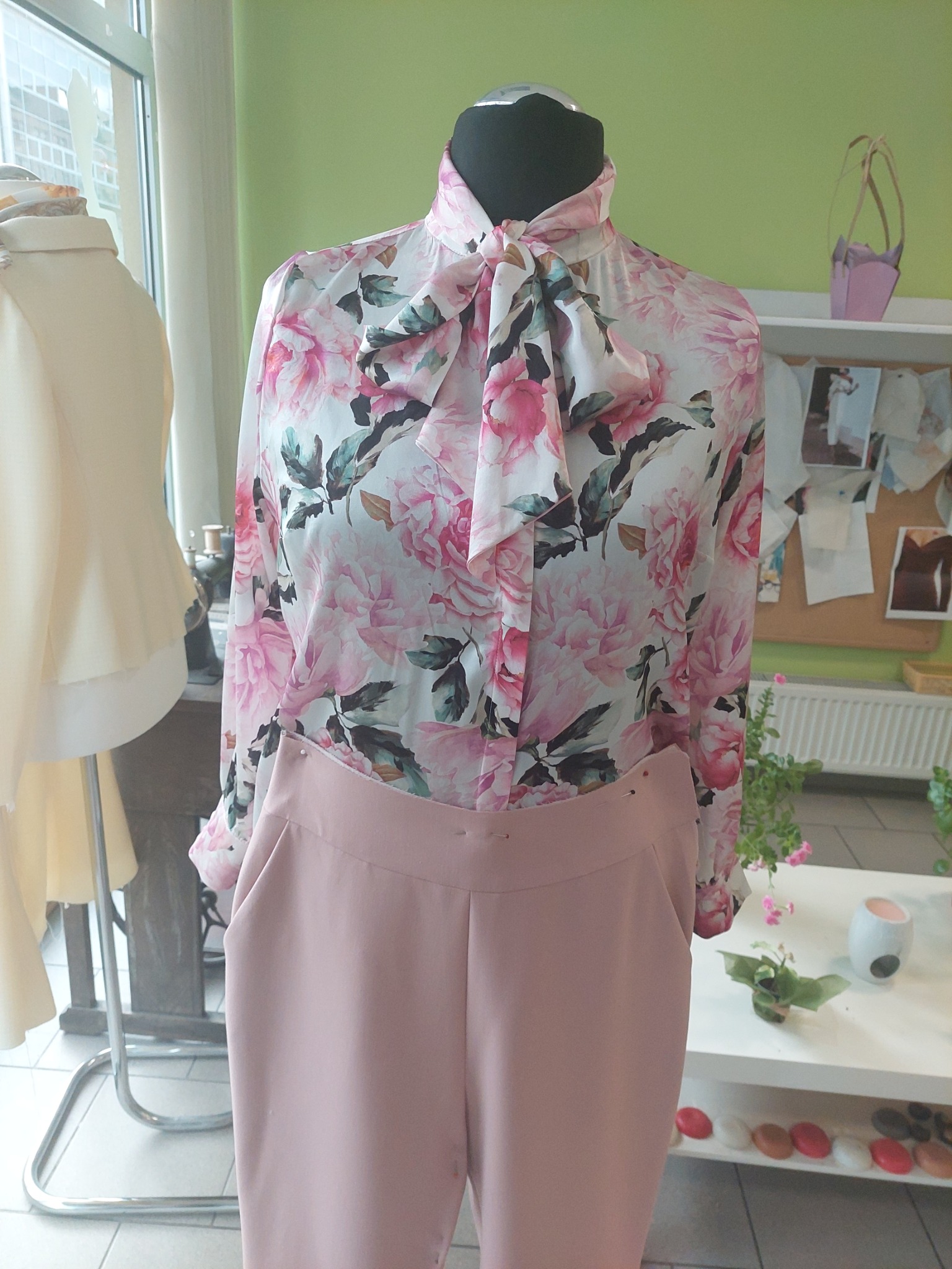 Manekin prezentuje bluzkę w różowe kwiaty z wiązaniem pod szyją oraz pudrowo-różowe spodnie. W tle widoczna pracownia krawiecka z akcesoriami.