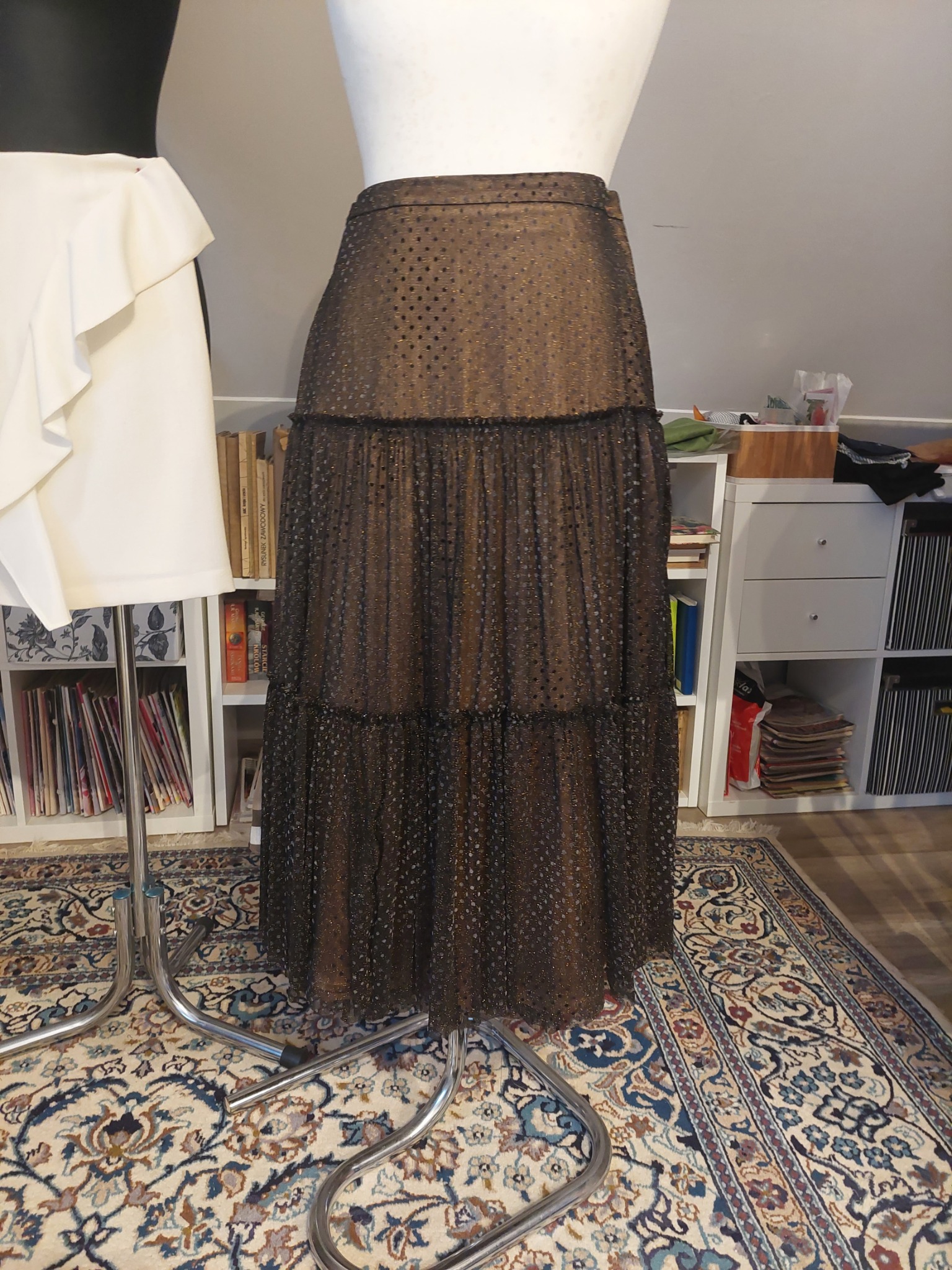 Brązowa, tiulowa spódnica z falbanami w kropki na manekinie krawieckim, na tle regałów z książkami i dywanu w orientalne wzory.