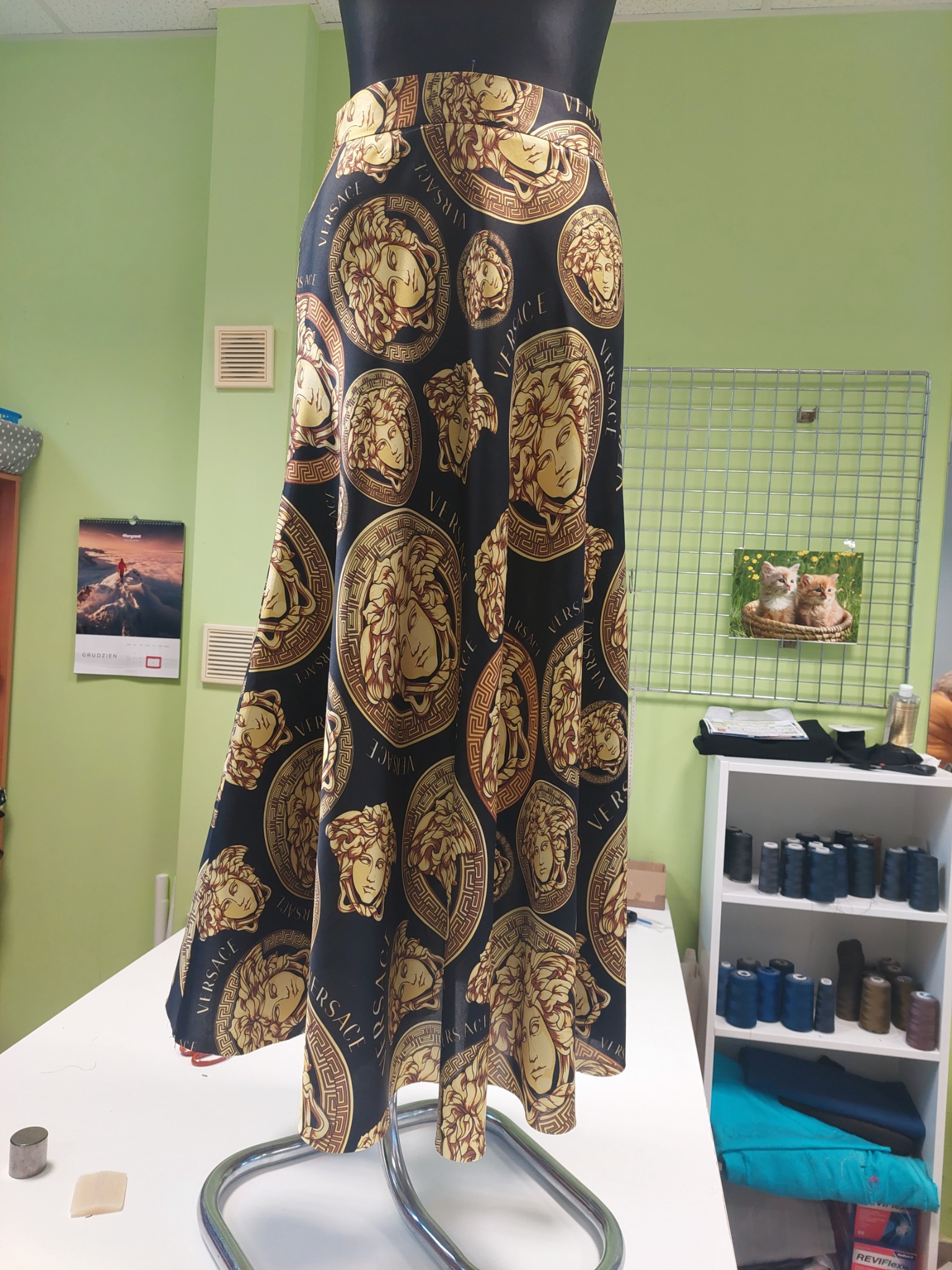 Długa spódnica z czarnego materiału w złote wzory Versace, prezentowana na manekinie w pracowni krawieckiej, z widoczną ścianą w kolorze seledynowym, kalendarzem i półką z nićmi.