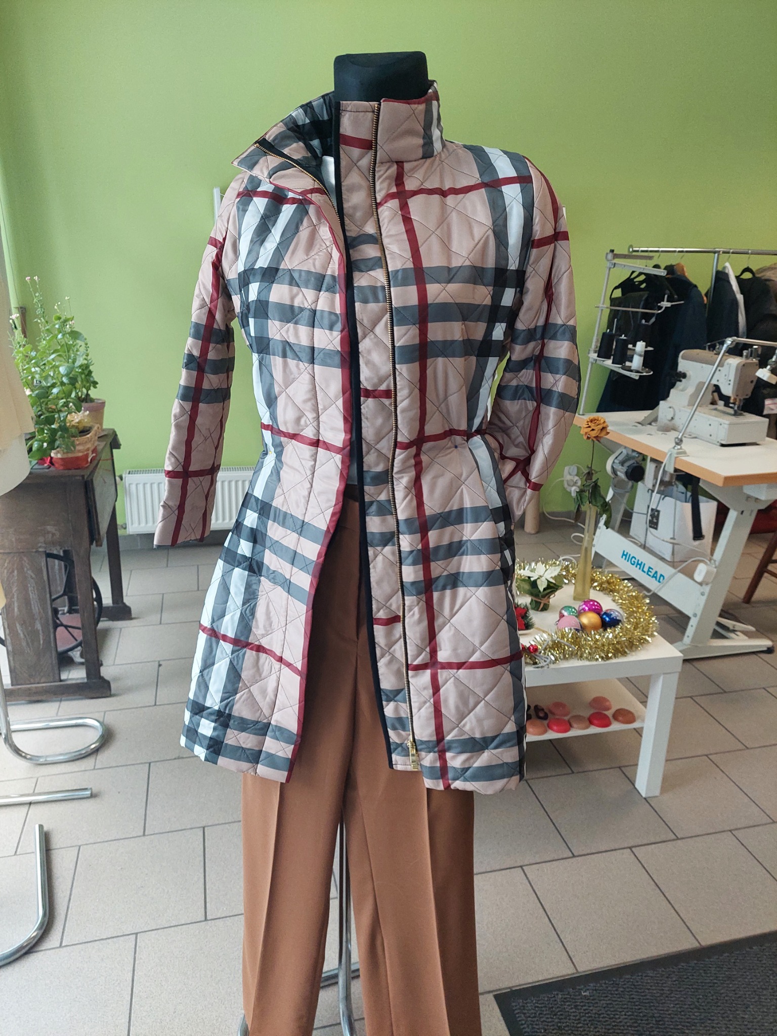 Manekin w rozpiętym, pikowanym płaszczu w kratę z brązowymi spodniami na tle pracowni krawieckiej z maszyną do szycia.
