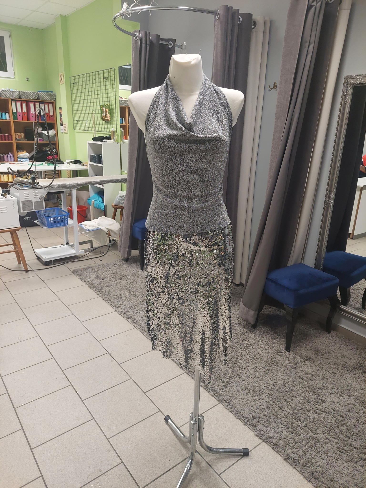 Srebrna, błyszcząca sukienka z drapowanym dekoltem i cekinową spódnicą na manekinie w pracowni krawieckiej, widoczne stanowisko do prasowania z parownicą i przymierzalnia z zasłonami w tle.