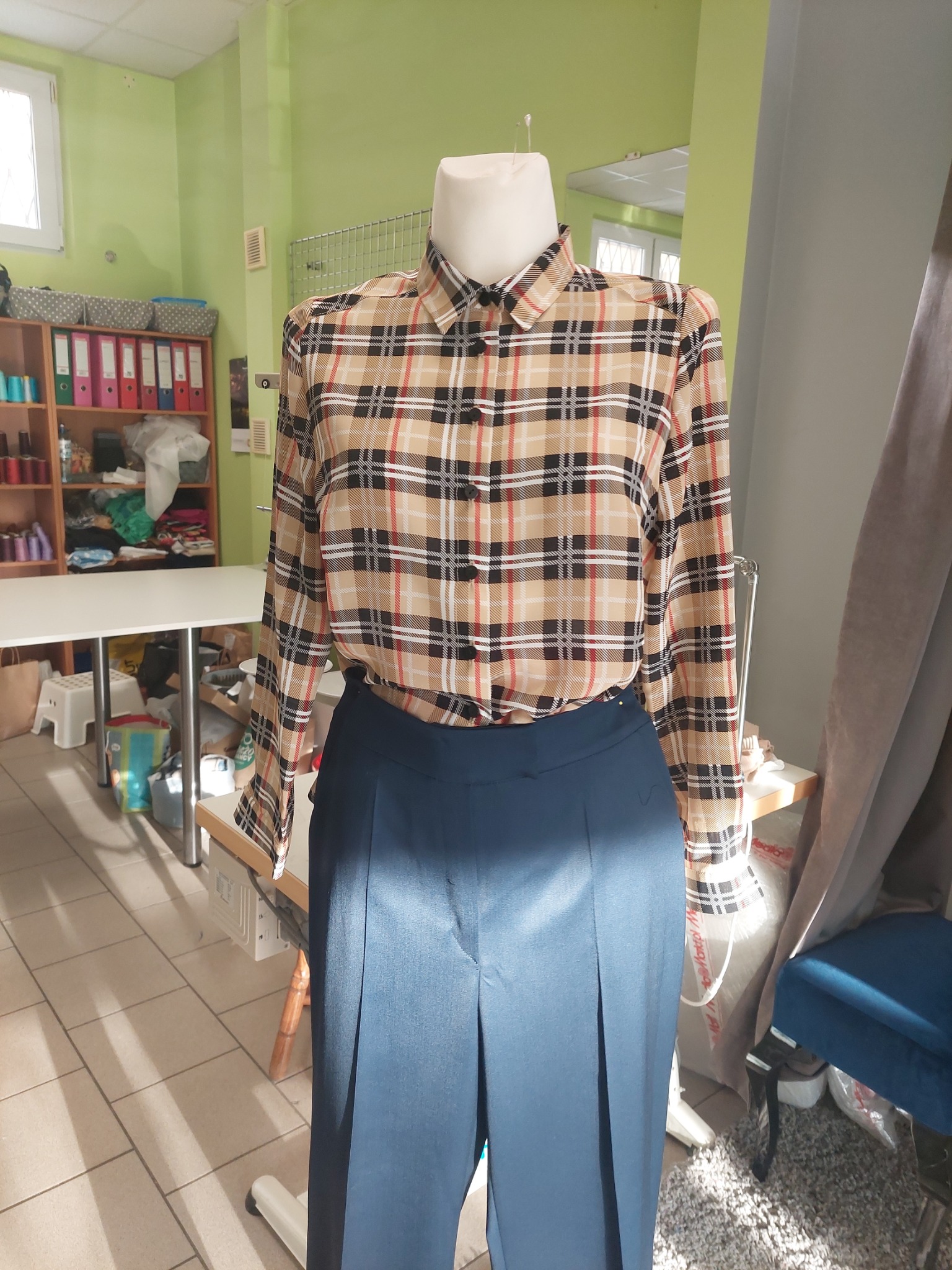 Manekin prezentuje bluzkę w kratę i granatowe spodnie w pracowni krawieckiej z zielonymi ścianami i regałem z materiałami w tle.