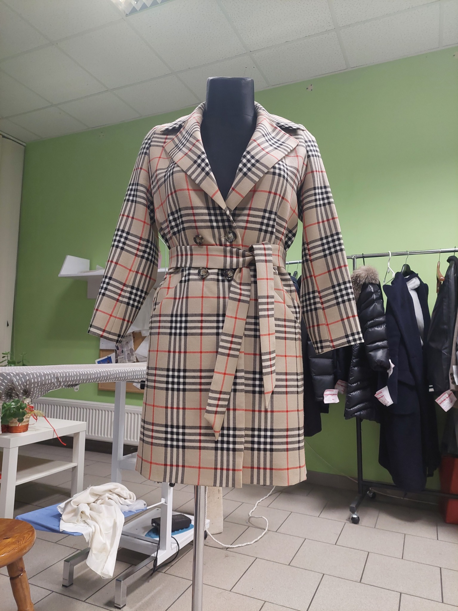 Beżowy płaszcz w kratę na manekinie krawieckim, z paskiem w talii, widoczny w pracowni krawieckiej z zieloną ścianą i wieszakami z ubraniami w tle.