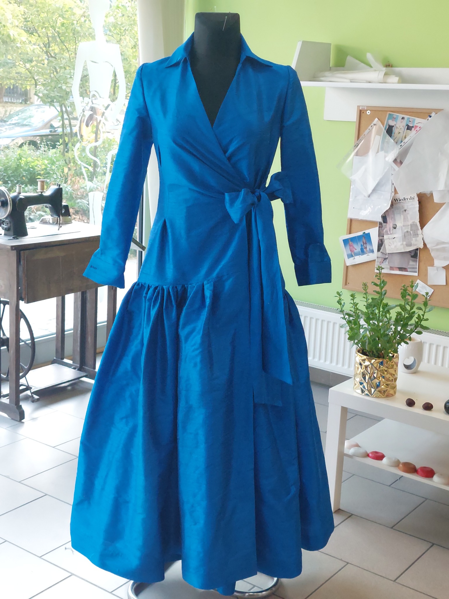 Gotowa, błękitna sukienka z wiązaniem w pasie, prezentowana na manekinie w pracowni krawieckiej, z maszyną do szycia Singer w tle.