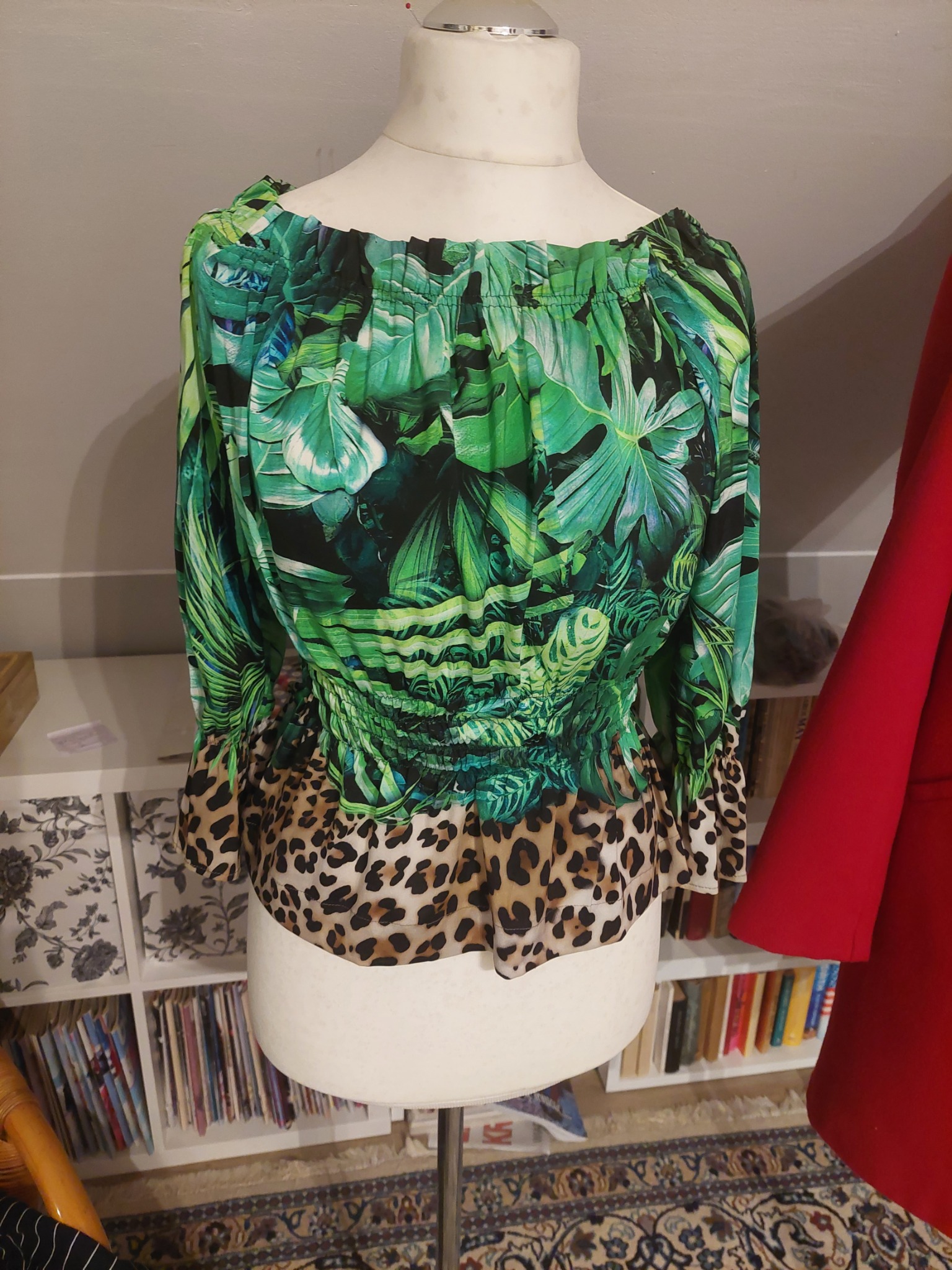 Bluzka z motywem tropikalnych liści i lampartem na manekinie krawieckim, na tle regału z książkami i czerwonej szaty.