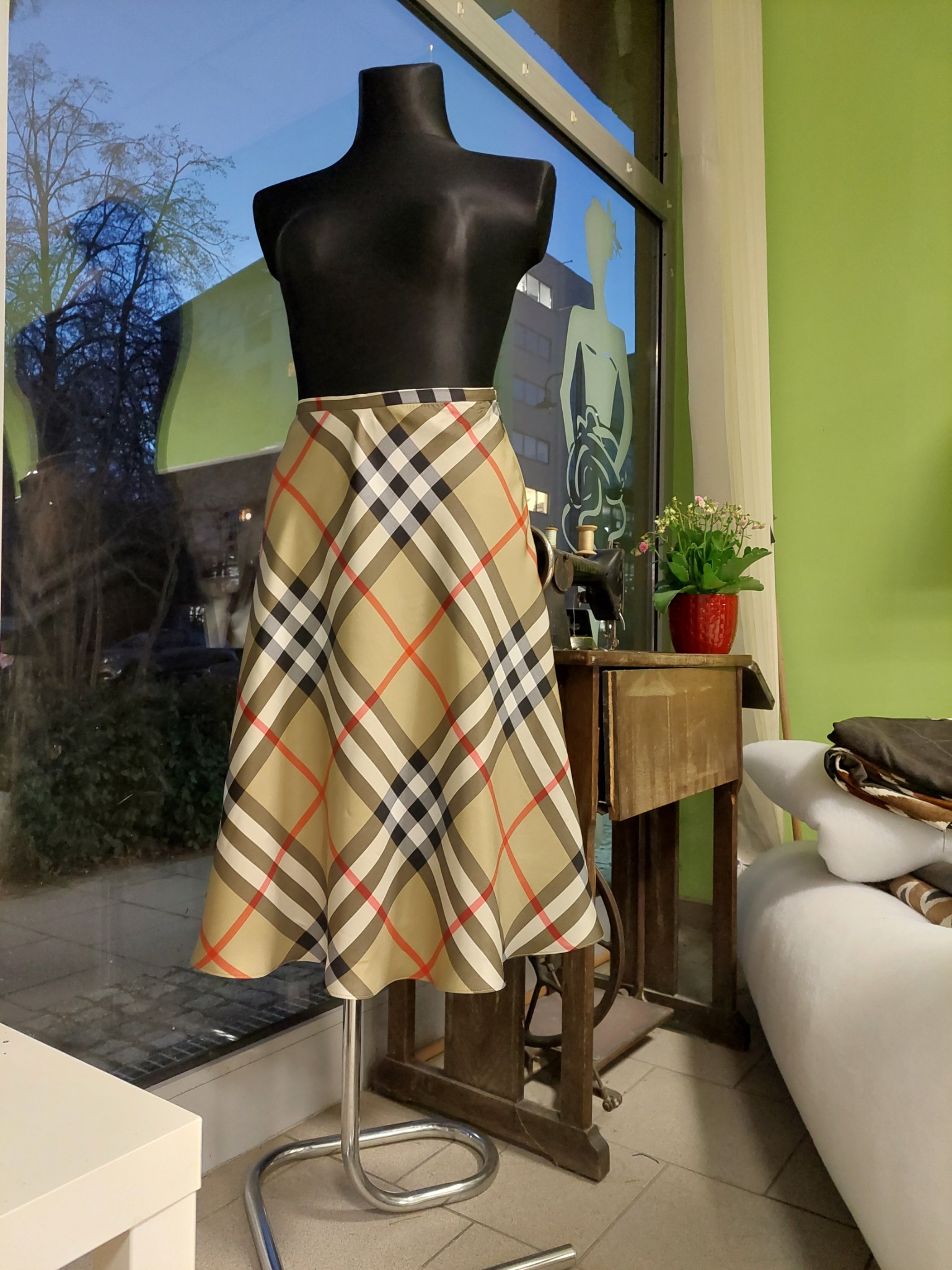 Spódnica w kratę na czarnym manekinie, stojąca przy oknie z widokiem na ulicę, obok zabytkowa maszyna do szycia i stos materiałów w pracowni krawieckiej.