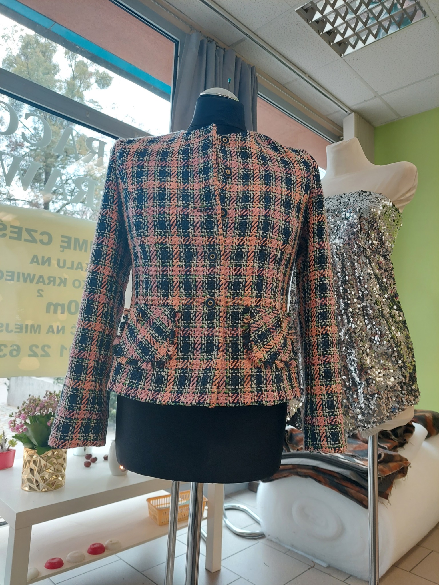 Tylny widok tweedowej marynarki w kratę w odcieniach różu i czerni, z ozdobnymi kieszeniami i guzikami, prezentowanej na manekinie w pracowni krawieckiej, obok sukienki cekinowej i kwiatów w złotym...
