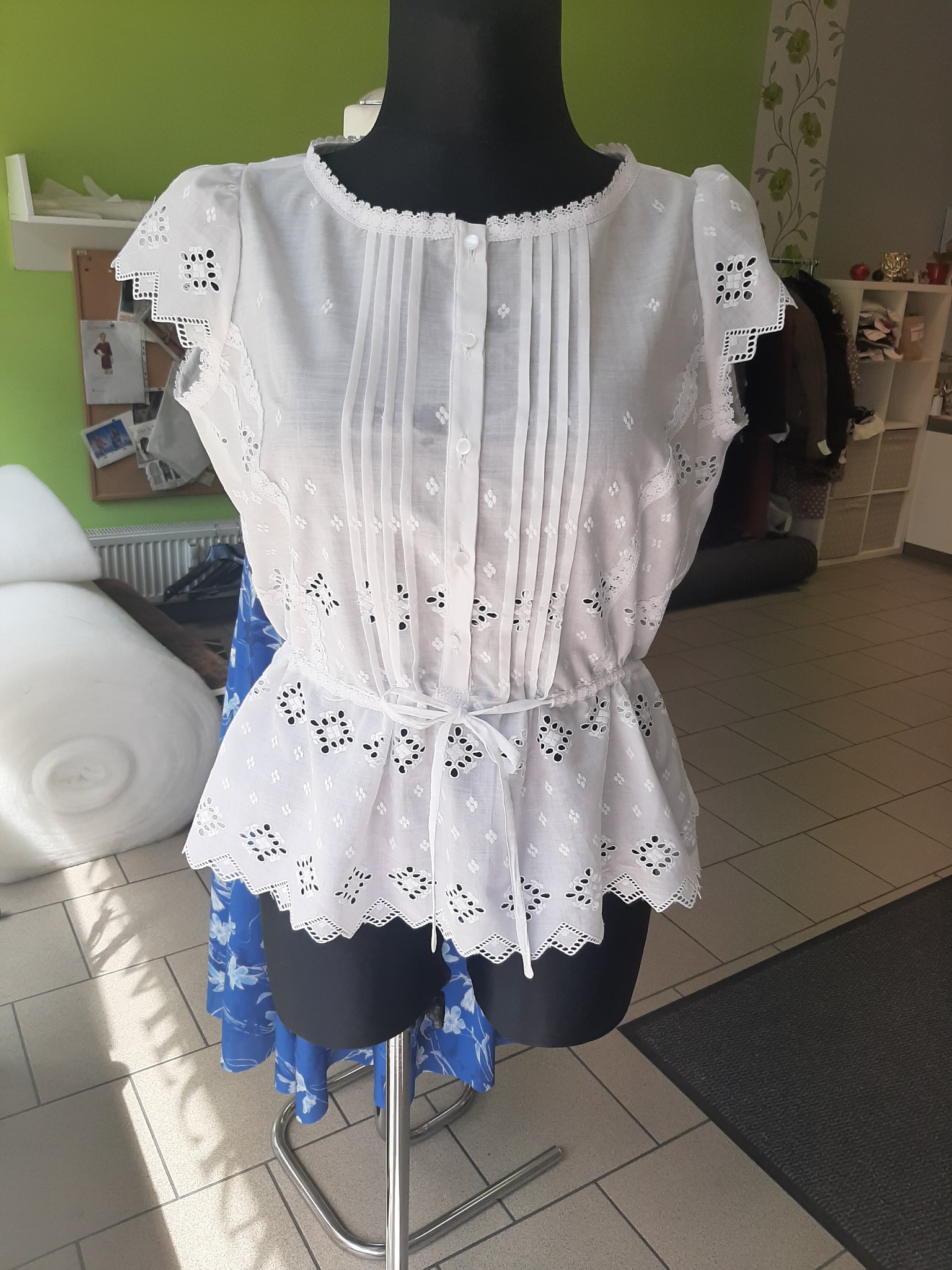 Biała bluzka z ażurowymi zdobieniami i plisami na manekinie krawieckim, z delikatnym paskiem w talii, prezentowana w pracowni krawieckiej.