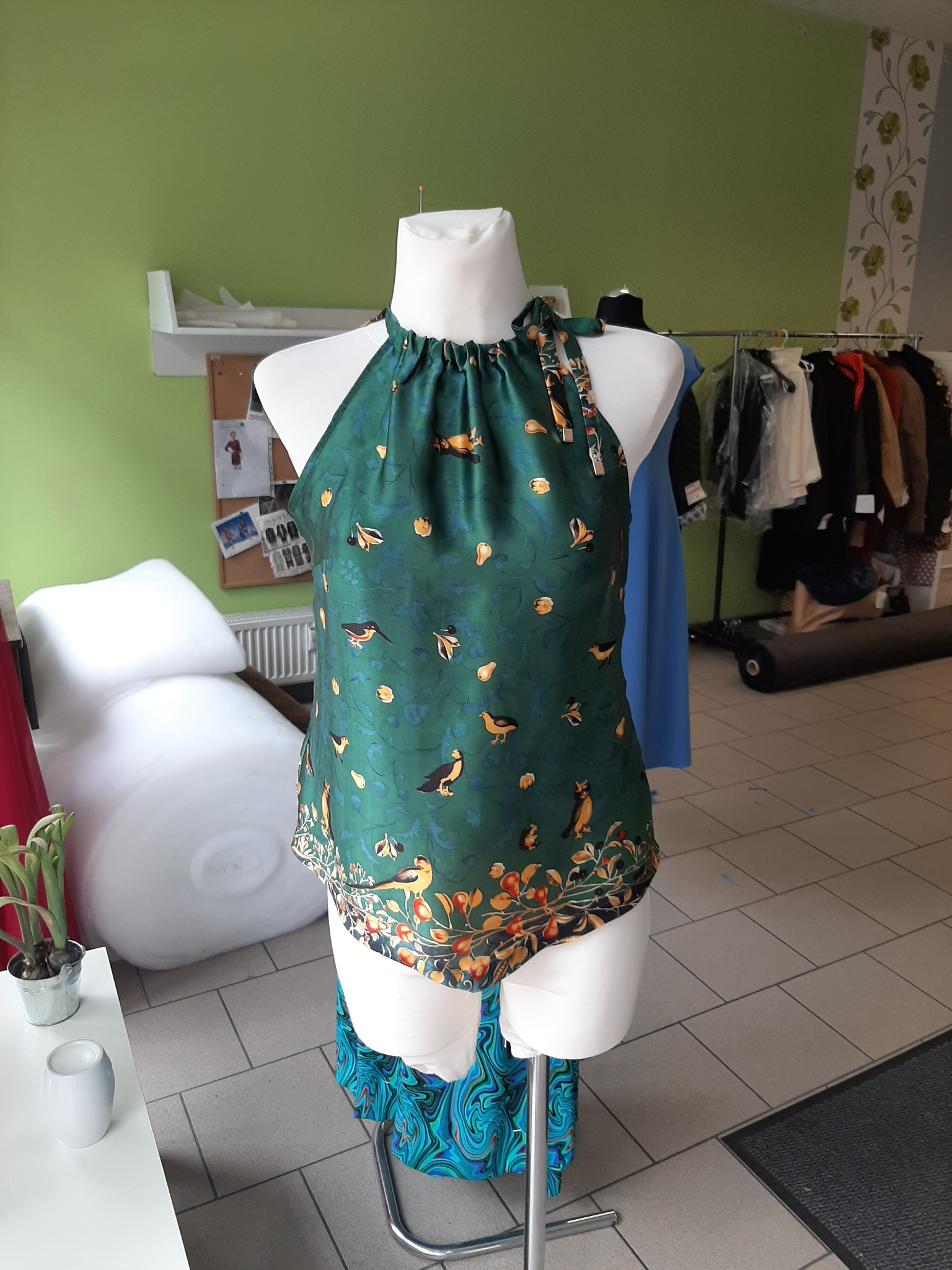 Zielona bluzka na manekinie z motywem ptaków i roślin, zawiązywana na szyi, widoczna w pracowni krawieckiej z rolką materiału w tle.