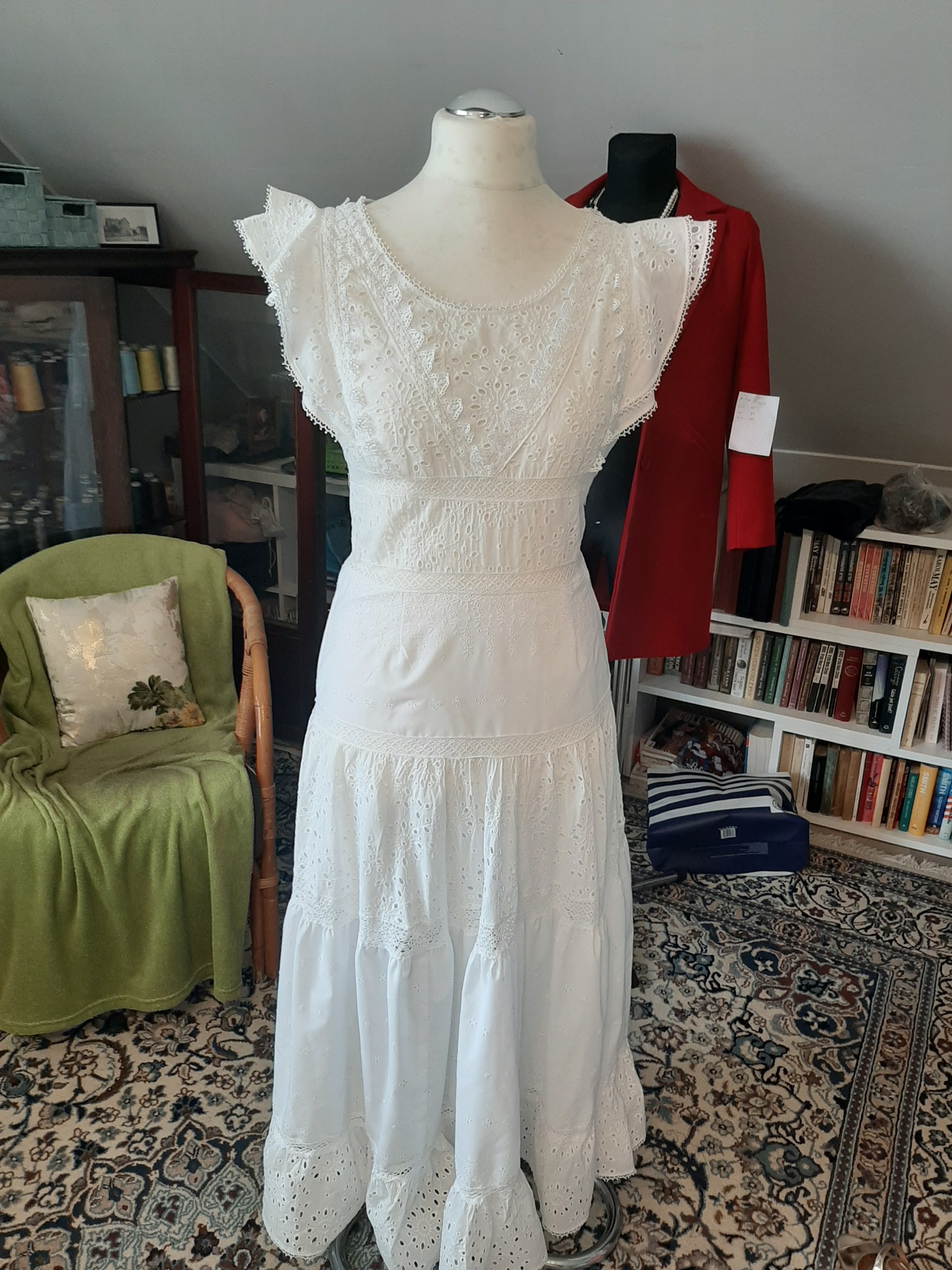 Biała, długa suknia z ażurowymi wstawkami, prezentowana na manekinie w domowym otoczeniu z dywanem w orientalne wzory, krzesłem z zielonym pledem i regałem z książkami w tle.