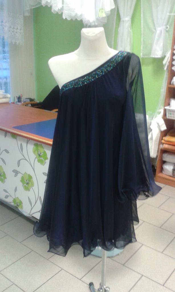 Granatowa, szyfonowa sukienka na jednym ramiączku, ozdobionym turkusowymi cekinami, zaprezentowana na manekinie w pracowni krawieckiej z zieloną ścianą i widocznymi firankami w tle.