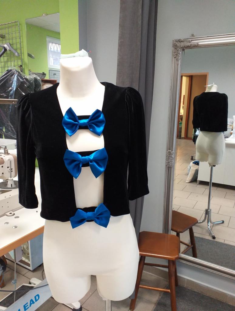 Czarny, welurowy żakiet z trzema, dużymi, błękitnymi kokardami na manekinie krawieckim, w tle widoczne lustro i maszyna do szycia.