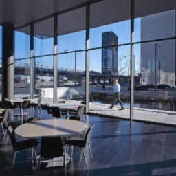 Wnętrze nowoczesnego biurowca z widokiem na miasto przez panoramiczne okna, ukazujące stoliki z krzesłami, przechodzącego mężczyznę i elementy infrastruktury miejskiej.