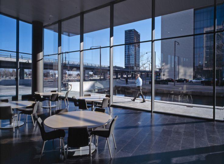 Wnętrze nowoczesnego biurowca z widokiem na miasto przez panoramiczne okna, ukazujące stoliki z krzesłami, przechodzącego mężczyznę i elementy infrastruktury miejskiej.
