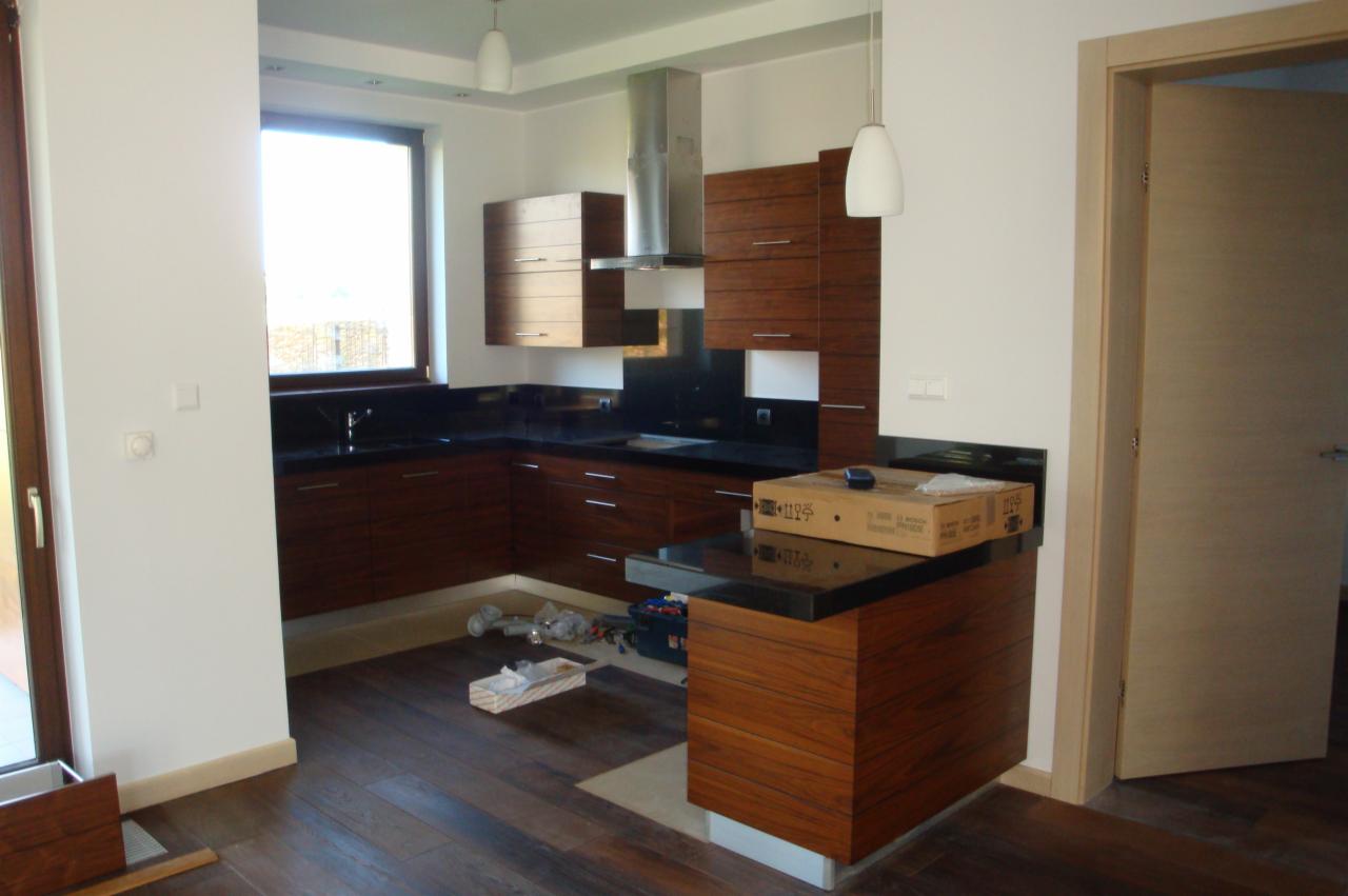 Montaż brązowych mebli kuchennych z czarnym blatem i okapem, widoczne kartony i narzędzia na podłodze, drewniana podłoga i białe ściany.