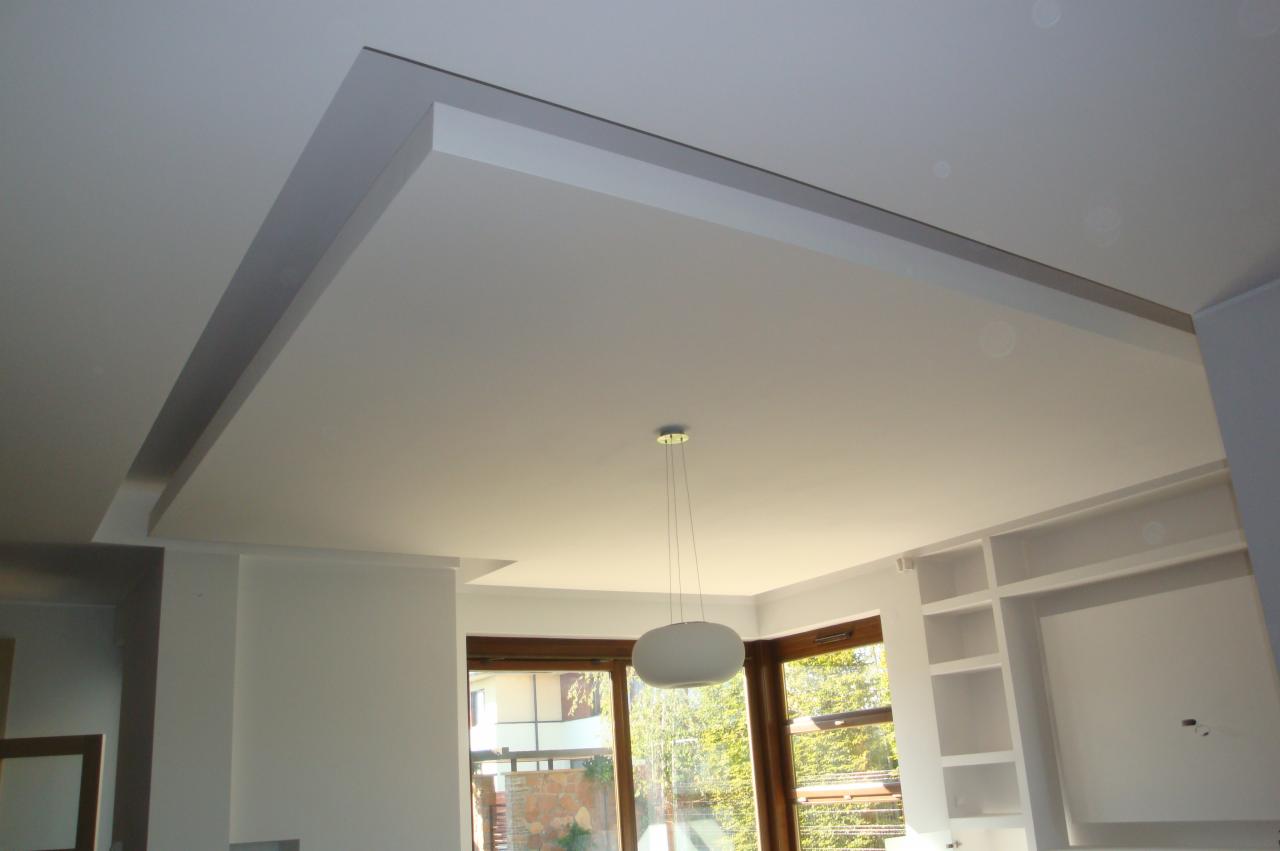 Nowoczesny sufit podwieszany z oświetleniem punktowym i dekoracyjną listwą, widok na okno i zabudowę meblową.