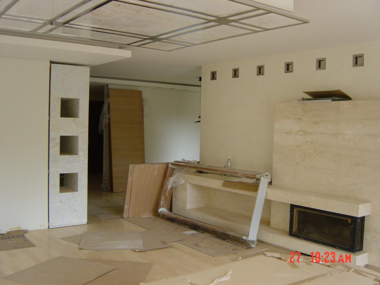 Wnętrze w trakcie wykańczania: marmurowa obudowa kominka, sufit z metalową kratownicą, fragment ściany z dekoracyjnymi wnękami, panele podłogowe i kartony zabezpieczające.