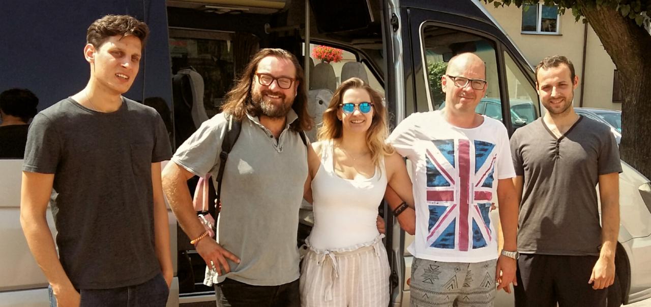 Pięcioosobowa grupa pozuje przed ciemnym vanem z kwiatami w oknie, uśmiechnięci, zrelaksowani, gotowi do występu.