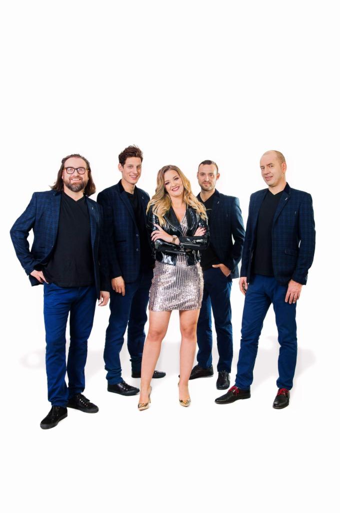 Pięcioosobowa formacja muzyczna pozuje na białym tle: wokalistka w srebrnej sukience i skórzanej kurtce na pierwszym planie, czterech mężczyzn w niebieskich marynarkach i spodniach w tle.