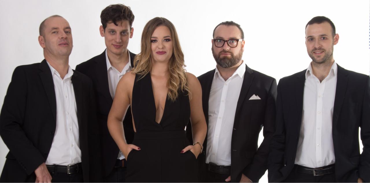 Pięcioosobowa grupa w strojach formalnych na białym tle: czterech mężczyzn w czarnych marynarkach i białe koszule, kobieta w czarnym kombinezonie z głębokim dekoltem, ręce w kieszeniach.